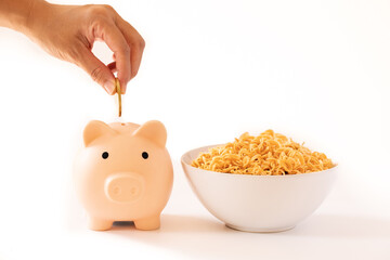 Close up hand putting coin in piggy bank beside instant noodles . in white bowl isolated on white background. Saving money or financial hardship concept. Saving money in daily life