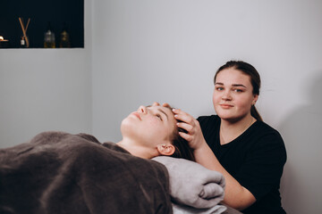 young beautiful spa customer laying down closed eyes covered with sliced organic cucumber on massaging bed while unrecognizable professional female masseuse therapist massage head and face