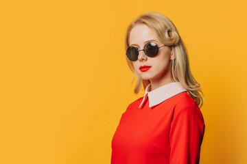 Stylish serious blonde hair woman in red dress and sunglasses on yellow background