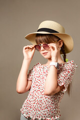 Portrait of stylish child girl in sunglasses, hat and summer wear posing at beige background, looking at camera. Pretty child lady with positive emotion. Fashion summer style concept. Copy text space