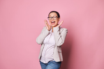 Surprised happy beautiful pregnant emotional woman, Latin American brunette expecting a baby, wearing trendy glasses, posing with open mouth, expressing amazement and joy, isolated on pink background