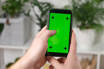 A man holds a phone with a green screen in his hands a background house plants. Mockup. Chromakey.
