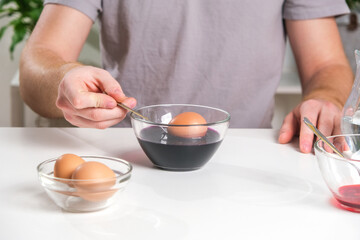 A man puts an egg on a spoon into a glass cup with a dye. Dyeing eggs with coloring in Easter egg