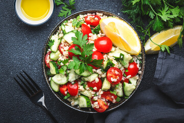 Bulgur tabbouleh salad with red cherry tomatoes, cucumbers, parsley and lemon dressing. Traditional Middle Eastern and Arabic dish. Bllack kitchen table background, top view