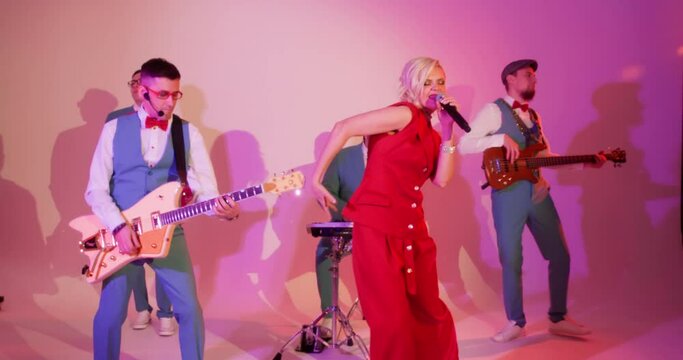 Music band woman singer and men musicians performing on stage in club. Artistic sensual woman in red singer emotionally sings song, men playing musical instruments and dancing. Live concert concept.