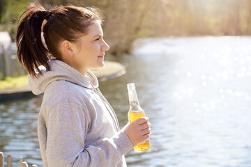 Woman on vacation drinking beer or lemonade from a bottle by a lake on a sunny day in summer