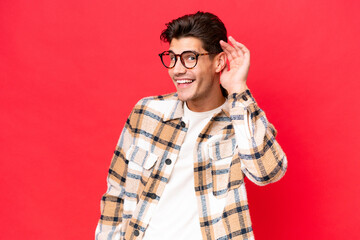 Fototapeta premium Young caucasian handsome man isolated on red background listening to something by putting hand on the ear