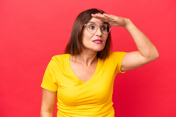 Middle-aged caucasian woman isolated on red background looking far away with hand to look something