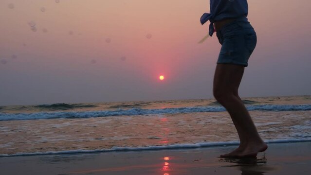 Silhouette of women's legs on the seashore against the sunset. Carefree woman enjoys her vacation and plays soap bubbles flying in the wind.