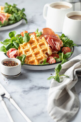 Cheesy belgian waffles served with ham, tomatoes and lettuce corn with cutlery on white marble background . Savory waffles. Healthy breakfast