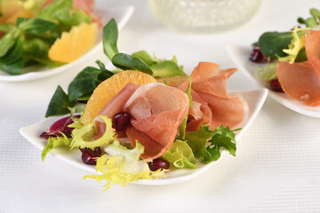 Portioned appetizer of prosciutto ham served on a of mixed with orange and pomegranate.