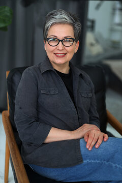 Portrait of a confident mature positive woman in glasses, sitting on an armchair in a home interior.