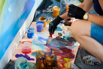 Artist hand in black gloves holds paint brush and choose color from colorful palette at outdoor art...