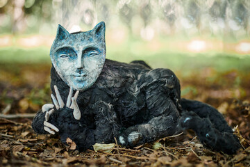 Black cat in human face mask art object at outdoor art exhibition in public park, weird black creature in mask lying on autumn foliage. Creepy strange cat art object, duplicity and fallacy concept