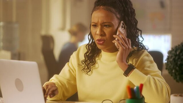 A young African American woman is typing on a laptop keyboard, talking on the mobile phone and very angry. A black female in a yellow sweatshirt sits at a table in the office. Close up.