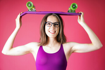 Cheerful happy skater girl with glasses wearing pink one piece swimsuit, posing with skateboard in...