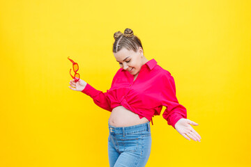 Happy pregnant woman with a smile on her face. Cheerful pretty pregnant woman in a pink shirt and pink glasses on a yellow background. Young bright pregnant woman.