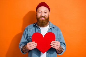 Photo of cheerful positive man wear jeans shirt smiling holding large red heart card isolated orange color background