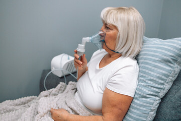 Sick upset caucasian senior woman with an inhaler. Unhealthy female doing inhalation at home, she use nebulizer and inhaler for the treatment, sitting on the couch at home, need treatment. 