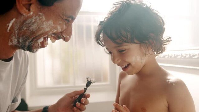 Dad and son laugh happily with shaving cream on their faces in a bathroom. This loving dad is teaching his son how to remove facial hair, a moment which turns into fun and enjoyment