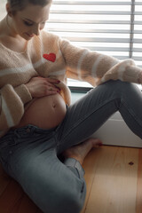 Charming young pregnant woman gently strokes and examines her stomach while sitting near window. Joyful emotions.