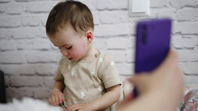 Adult’s hand showing a phone to a little toddler. Baby looks at gadget with interest. White wall at backdrop.