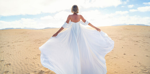 Back view of a blonde woman in fashionable, maxi white wedding dress posing on the desert, dancing...