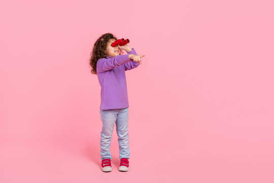 Full length photo of excited little child dressed purple pullover watching binoculars pointing empty space isolated pink color background