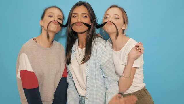 Three young beautiful smiling hipster girls in summer clothes. Sexy carefree women having fun.Positive models going crazy near blue wall in studio.They make moustache with hair
