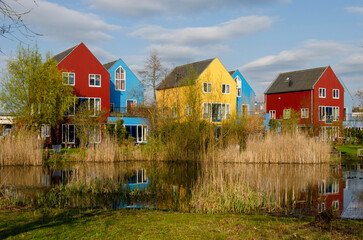 Fototapeta premium Culemborg, The Netherlands, April 8, 2023: Scandinavian style houses with wooden facades in primary colors reflect in a pond in the golden hour on an evening in spring