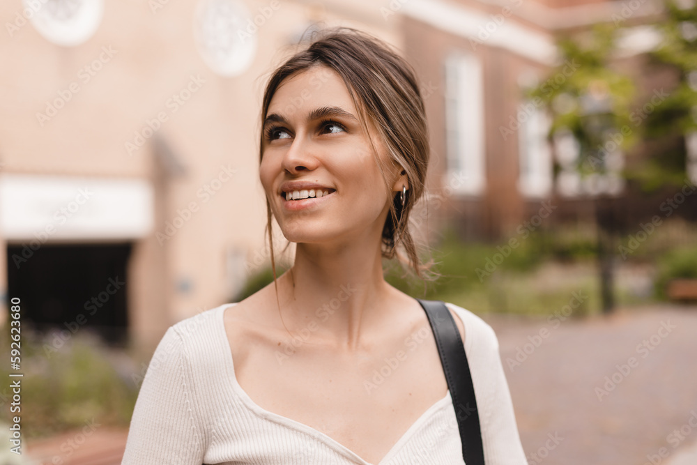 Wall mural Close-up of young caucasian woman looking seductively into side and smiling. Beauty with low bun hair, wears top and bag on shoulder.