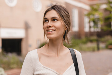 Close-up of young caucasian woman looking seductively into side and smiling. Beauty with low bun hair, wears top and bag on shoulder.