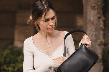 Fototapeta na wymiar Elegant young woman looking in her black leather bag her phone or purse. Business style woman wear long sleeve top, low bun hairstyle and bag on the street. Street style, fashion outfit.