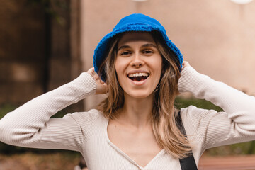  Pretty young caucasian woman look at camera, hold hat panama, spends her leisure time on walk in street. Brunette wears blue bright panama hat, top with long sleeves. Positive emotions concept.