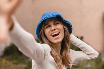 Close up young caucasian woman look at camera, hold her panama hat, spends her leisure time on walk in street. Brunette wears blue panama hat, top with long sleeves. Girl make selfie photo, open mouth