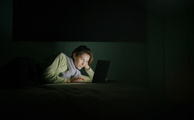 A young woman is lying on her bed with a laptop, working or studying through the night. The soft light of the laptop screen illuminates her face in a dark room