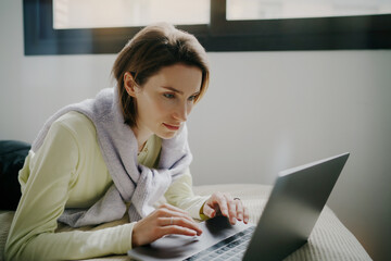 Young womanstudying hard on her laptop, while lying comfortably on her bed in a sunny room, taking a break from work to enjoy a movie or browse the internet.