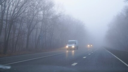 A car with its headlights on is driving in the fog along on an asphalt road