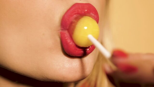 Closeup red lips of young beautiful hipster bad girl with earring in her nose. Sexy carefree smiling woman posing in studio near beige wall. Positive model licking round candy lollipop