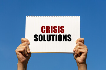Crisis solutions text on notebook paper held by 2 hands with isolated blue sky background. This message can be used as business concept about crisis.