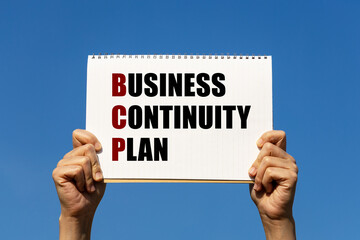 Business continuity plan text on notebook paper held by 2 hands with isolated blue sky background. This message can be used as business concept about BCP.