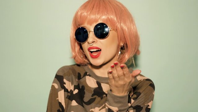 Portrait of young beautiful hipster bad girl in trendy summer clothes and earring in her nose. Sexy carefree smiling woman in studio in pink wig near wall. Positive model gives air kiss