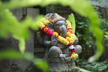 Traditional Ganesha statue with flower wreath in Balinese garden. Lord Ganesha is believed to bring...