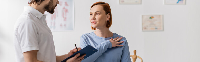 woman touching injured shoulder near physiotherapist writing diagnosis on clipboard in consulting...