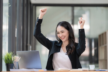 Successful young Asian business woman achieving goals excited raised hands rejoicing with laptop in the office.