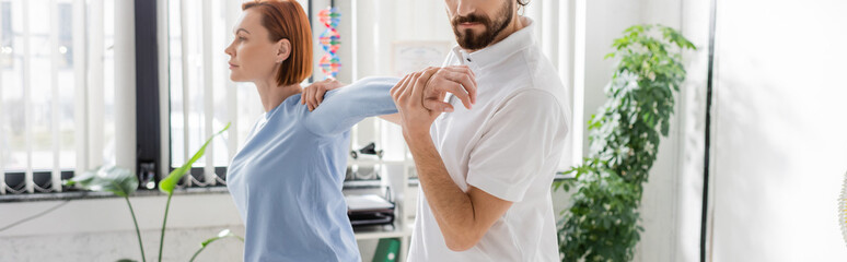 bearded physiotherapist stretching arm of redhead woman while doing diagnostics in consulting room,...