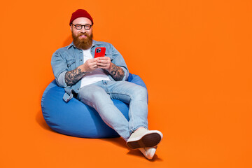 Full length photo of funky cool guy dressed denim jacket relaxing bean bag typing gadget empty space isolated orange color background