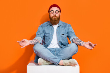 Photo of dreamy funky guy dressed denim jacket closed eyes enjoying yoga sitting cube isolated orange color background