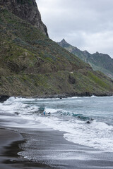 People surfing alone in a black sand beach with waves surrounded by nature and mountains in cloudy day in tenerife, canarias, spain