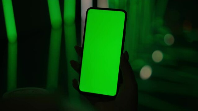 Mobile phone in hand. Holding smartphone with red blue neon lighting on dark background. Green chromakey screen. 4K